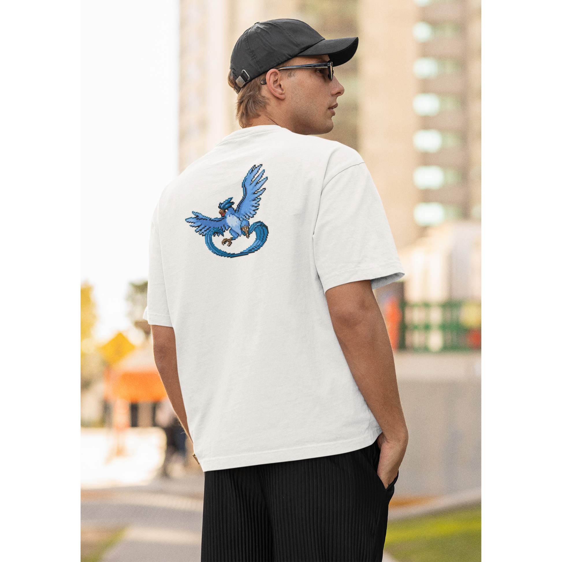 A man with short brown hair and a hat is standing on a busy city street, wearing a white T-shirt with a Pokemon  print. He is facing away from the camera, showcasing the back of his white T-shirt, which features a detailed pixel art design of Articuno, the legendary ice Pokémon.