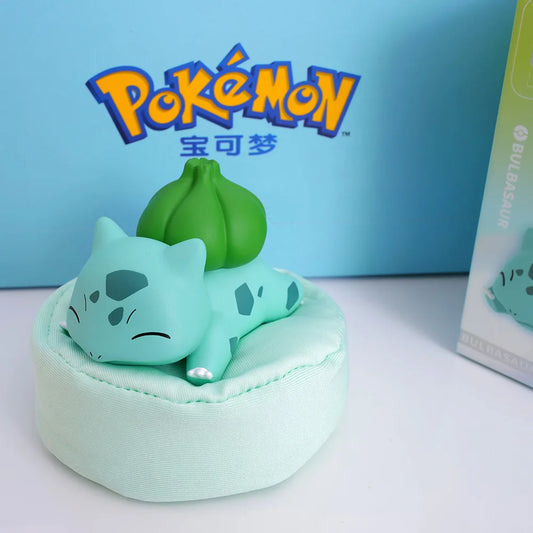 Bulbasaur figure sleeping on a pastel green cushion/pillow, featuring a cute design inspired by Pokémon, with a branded Pokémon box in the background.
