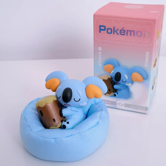 Komala figurine sleeping on a blue cushion/pillow, holding its signature log, with a matching Pokémon-themed box in the background.