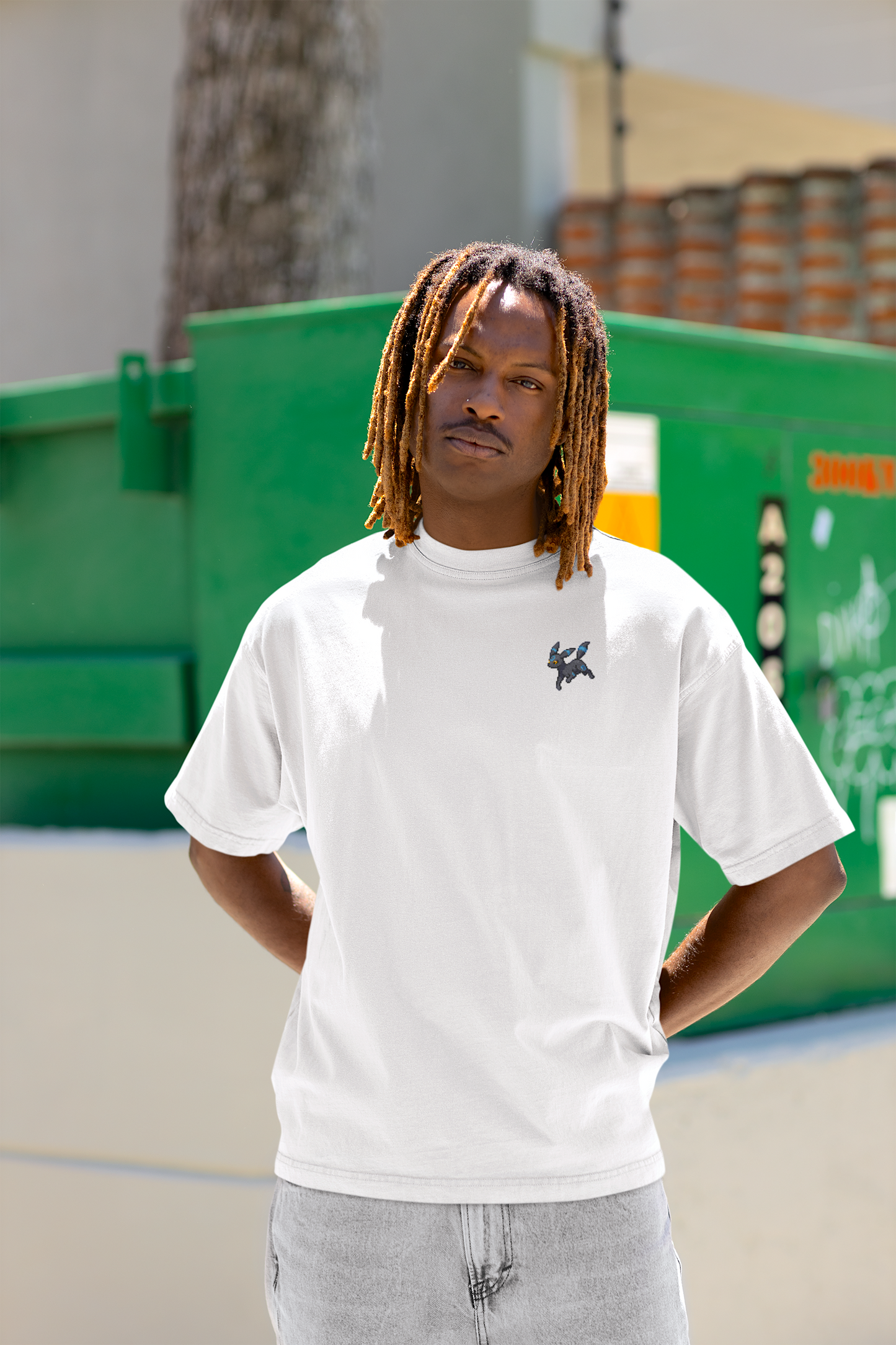 A young adult black male model with a relaxed demeanor is captured in a candid shot, wearing a white T-shirt with a vibrant Shiny Umbreon Pokemon pixel art design. The model's outfit is casual and stylish streetwear look.